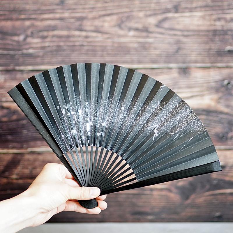 箔図 黒骨扇子7寸を手に持ち広げた正面——黒地に銀箔が舞う京扇子｜米原康人